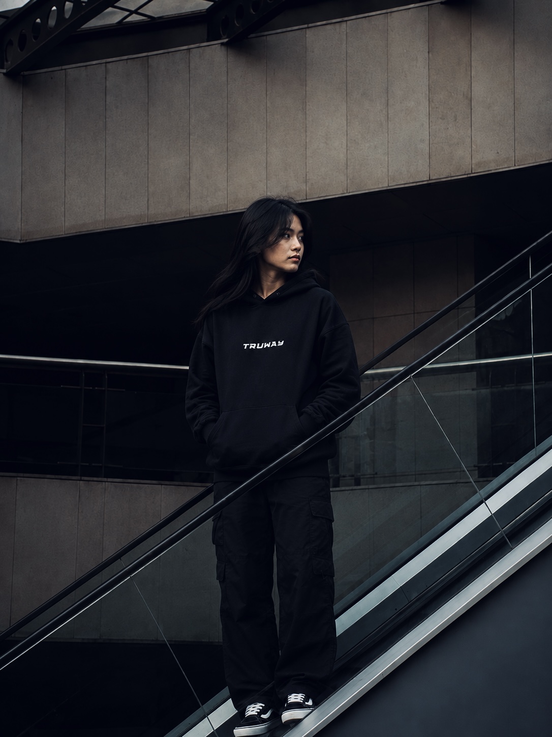 A woman in the black TRUWAY hoodie on an escalator in a concrete building